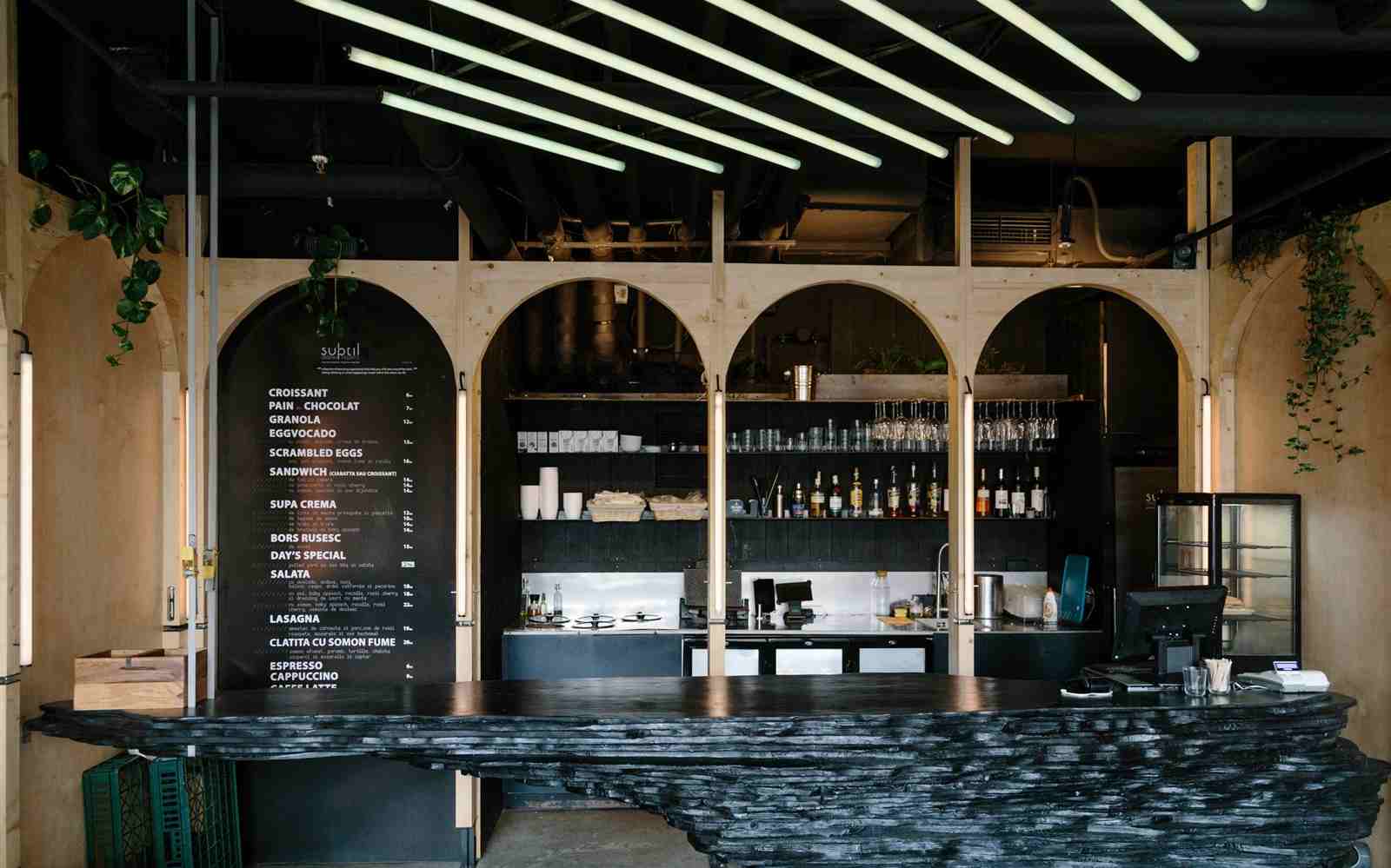 Bar negru cu formă neregulată din lemn ars și rafturi pentru produse în spatele unor arcade din lemn