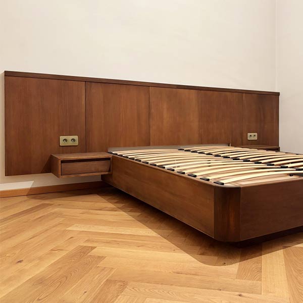 Pat din lemn masiv la nuanta nucului, cu colturi rotunjite, cu tablie formata din panouri decorative aplicate pe de perete, si noptiere suspendate