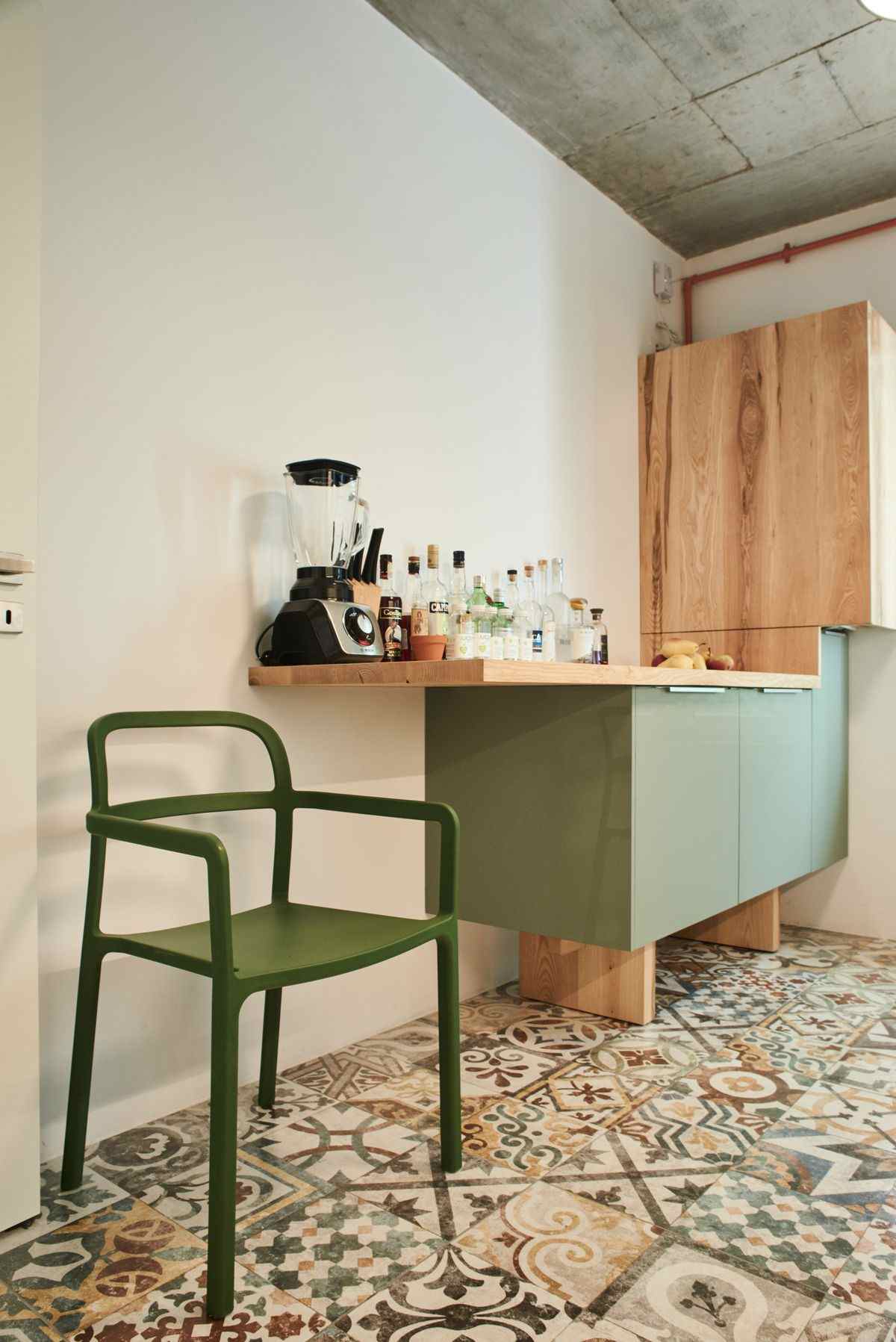 Corp de bucătărie depărtat de pardoseala cu picioare masive din lemn. Dulapurile au o nuanță verde oliv, iar blatul de bucătărie se continua in consola. In plan secund este o masca pentru centrala termica din lemn de frasin cu aspect natural.