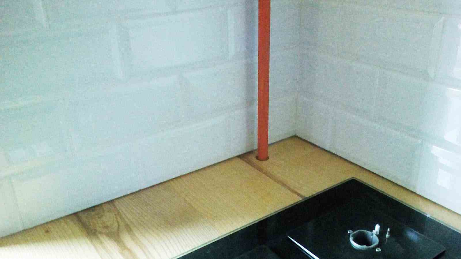 Detaliu de la coltul blatului de bucatarie din lemn cu teava portocalie ce patrunde prin blat, langa plita. Peretele este placat cu o faianta alba in relief.