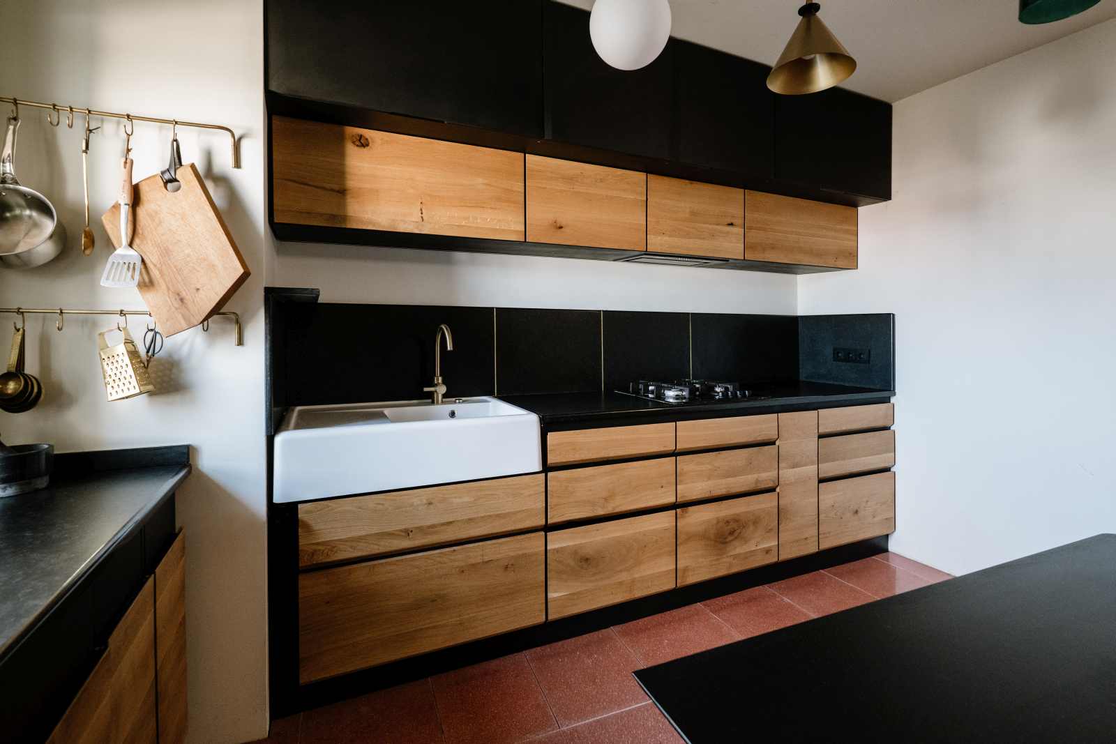 Imagine de ansamblu cu interior de bucatarie in combinatie de placi negre si fete de sertare si usi din lemn de stejar, pe doua laturi langa fereastra cu dulapuri joase si corpuri suspendate sus, chiuveta ceramica alba, rastel de ustensile pe perete